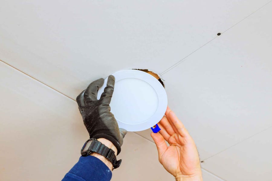 An electrician is carefully placing LED light fixture into ceiling opening during home renovation.